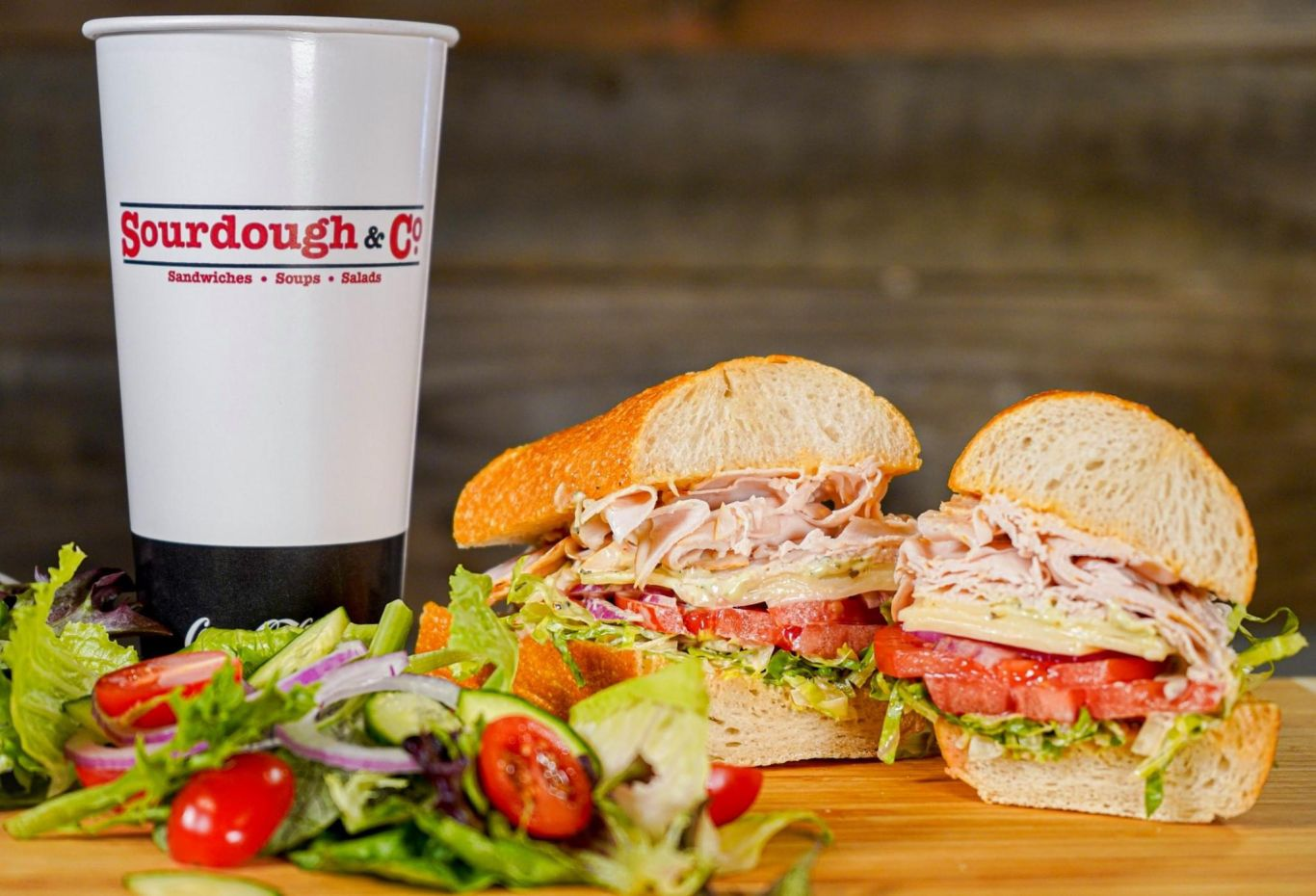 A sandwich from Eureka Sourdough & Co. in California with turkey, lettuce, tomato, and cheese on sourdough bread is next to a cup labeled "Sourdough & Co." A side salad of mixed greens, tomatoes, and red onion completes this tasty meal. Perfectly balancing our offerings of sandwiches and fresh soups & salads.