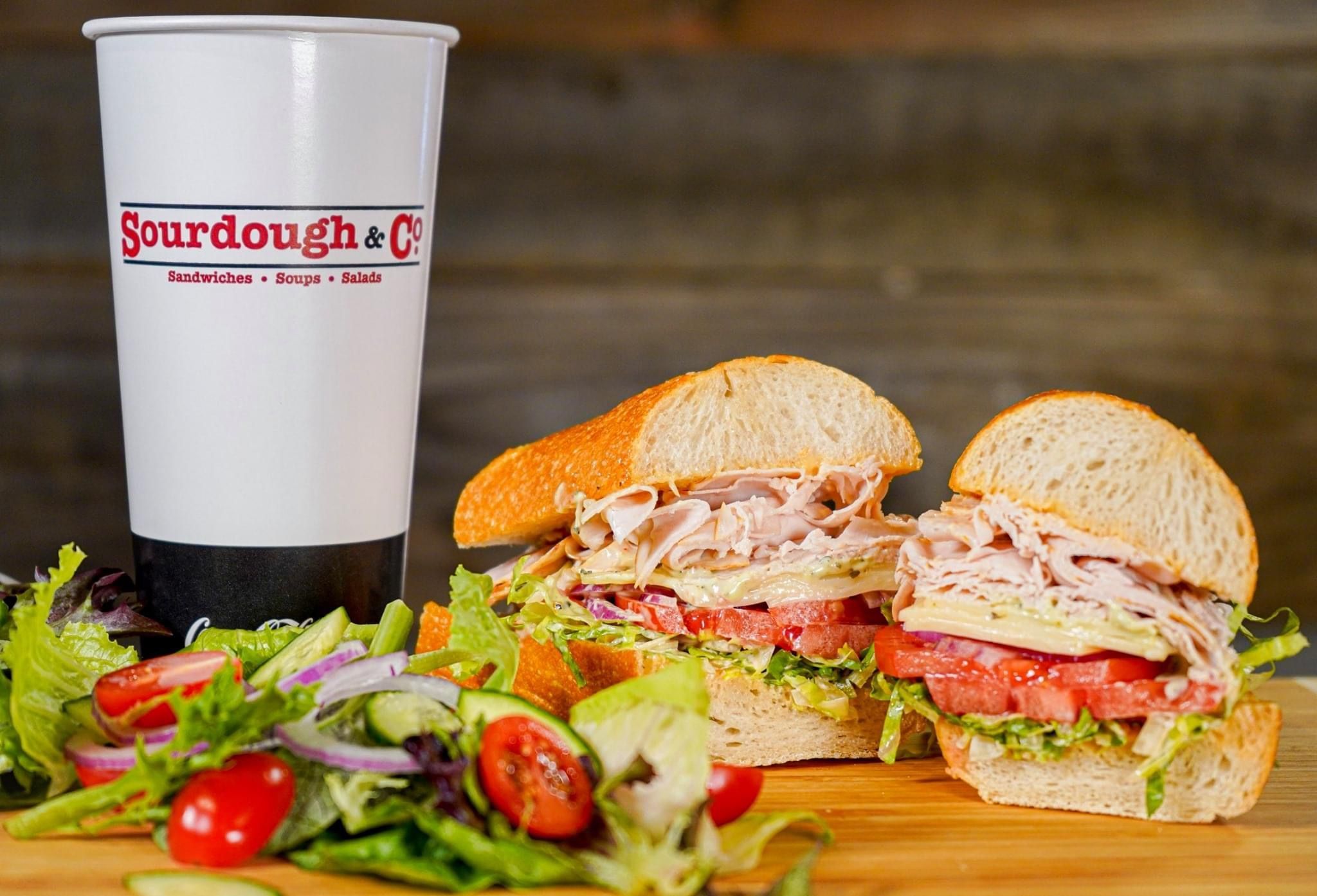 A sandwich from Fairfield North Texas St Sourdough & Co. in California with turkey, lettuce, tomato, and cheese on sourdough bread is next to a cup labeled "Sourdough & Co." A side salad of mixed greens, tomatoes, and red onion completes this tasty meal. Perfectly balancing our offerings of sandwiches and fresh soups & salads.